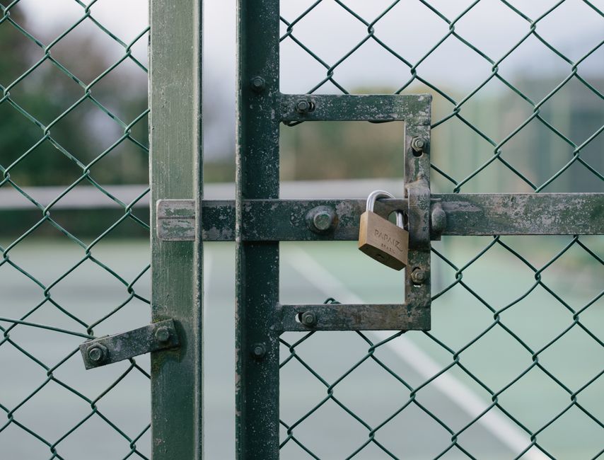 A close-up view of a weathered green metal chain-link fence with a rectangular latch mechanism secured by a small padlock engraved with 'PITMAN,' set against a blurred outdoor tennis court background.