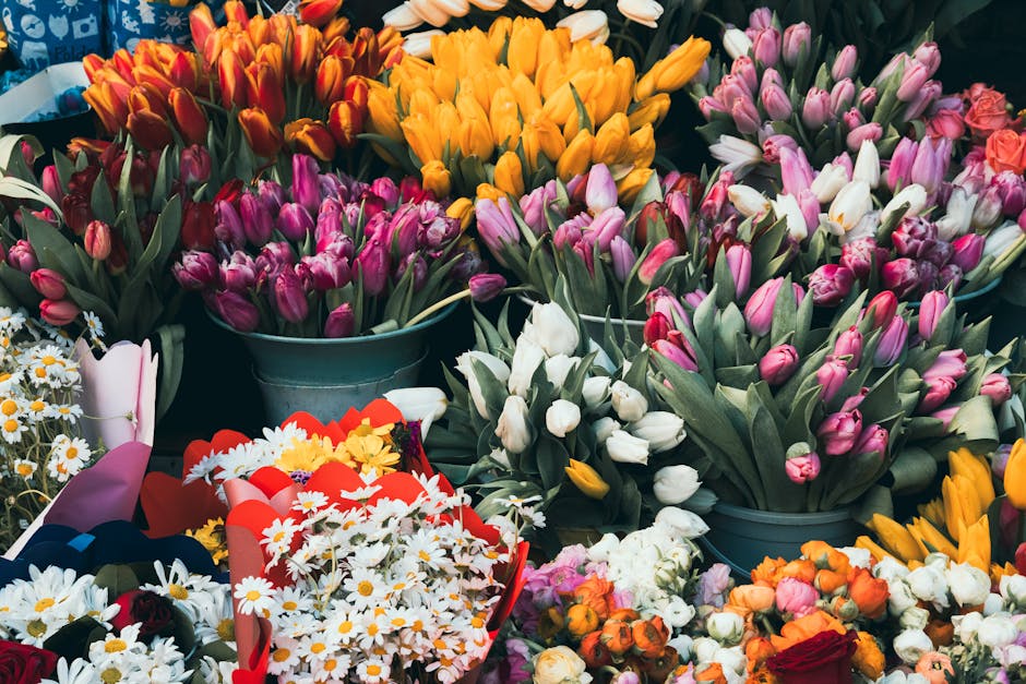 A vibrant display of fresh floral arrangements featuring a variety of tulips in shades of pink, white, orange, and yellow, arranged in metal buckets and baskets. The tulips have tightly closed buds an