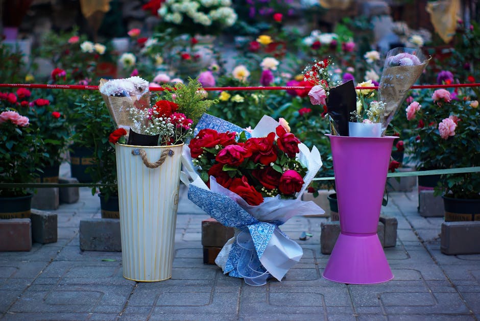 A floral arrangement featuring a bouquet of vibrant red roses wrapped in white and light blue paper, placed on a paved area outdoors. Flanking the bouquet are two tall vases: a cream-colored striped v