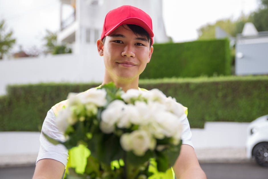 A delivery person wearing a red cap and a neon yellow safety vest stands outdoors, smiling as they extend a bouquet of fresh white flowers towards the camera. The floral arrangement features roses and