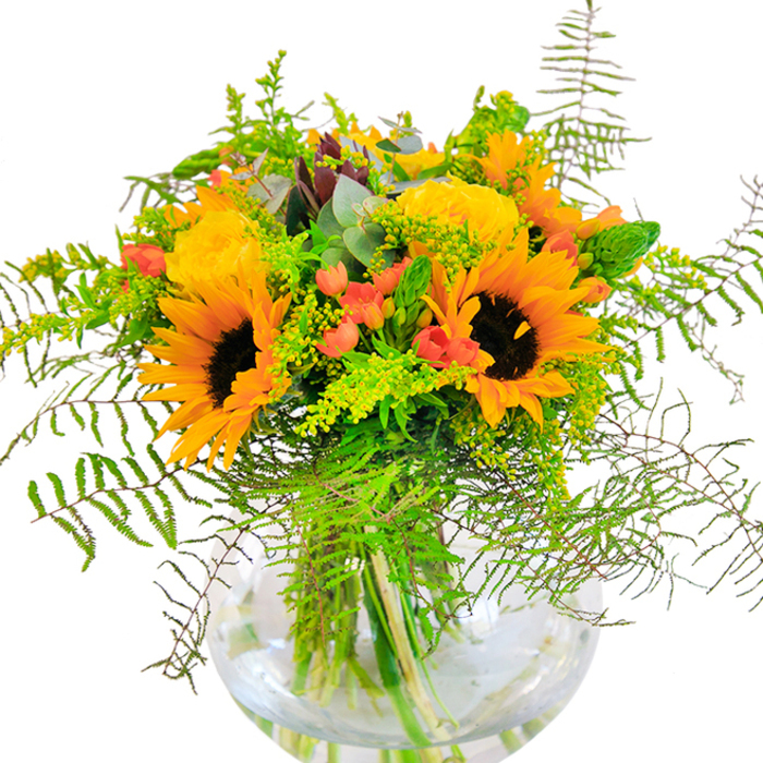 A radiant celebration of Charlton's local charm, this striking floral arrangement captures the warmth of golden afternoons by the Thames and the vibrant atmosphere of Woolwich Road's bustling markets. Generous sunflowers, their brown-black centers ringed with sunbeam petals, anchor the display, each bloom a small sun in its own right. Tucked among them are buttery yellow garden roses, their tightly coiled centers and velvety petals creating harmonious contrast and visual intrigue. Glossy, crimson Hypericum berries are scattered with care, bringing playful pops of colour that animate the sea of yellow and green. Airy fern fronds and fine green filler cascade outward with gentle motion, mimicking the greenery found along Charlton's walking paths and leafy promenades. Hints of eucalyptus, light blue-green and aromatic, offer calming undertones to the cheerful composition. The round glass vase not only provides a modern finishing touch but also showcases the tangle of bright stems as they crisscross in clear water-an image of health and vitality. Whether chosen as a gift for new parents, to mark an anniversary, or simply to add a spark of joy to a local gathering, this beautifully balanced bouquet brings together the season's best for delivery across Charlton and its surroundings, crafted with care and a florist's passion for the neighbourhood's natural beauty.