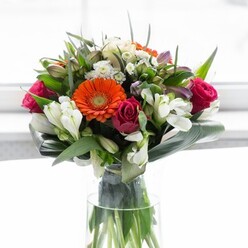 This vibrant bouquet, artfully composed by the skilled hands of Charlton's local florists, celebrates the delicate interplay of colour, structure, and sentiment. Creamy gerbera daisies with glowing orange hearts form a luminous crown, their gentle petals radiating optimism in a scene reminiscent of sunlight dappling through Maryon Wilson Park. Deep fuchsia roses add rich, velvety layers, hinting at romance and the joy of connection. Creamy white alstroemeria and petite white blossoms are scattered throughout, evoking soft clouds drifting past the Royal Arsenal Riverside. Broad, glossy leaves-artfully wrapped around the base-ground the lively colours, their verdant greens rooted firmly in tradition, like the trees lining Charlton Park. Additional slender greenery and textured sprigs fill the gaps, inviting the viewer to appreciate each bloom's uniqueness. The bouquet sits in a sleek, transparent vase, with sturdy stems visible, reinforcing its freshness and meticulous preparation. Bathed in diffuse daylight, each flower gleams, while the overall arrangement feels balanced, rejuvenating, and full of possibility. The subtle mingling of floral fragrances conjures a sense of new beginnings, making this display ideal for welcoming friends, gathering family, or offering encouragement in Charlton's ever-welcoming community.